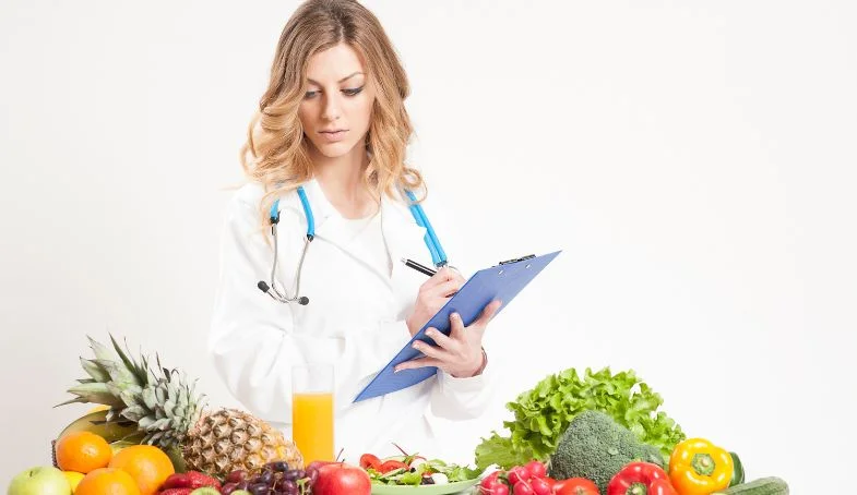 Principios Básicos de la Nutrición. Doctora Nutricionista tomando notas mirando una mesa llena de verduras, frutas y vegetales
